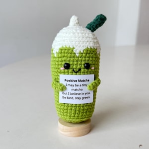 Könnte beinhalten: Ein gehäkeltes Matcha-Getränk mit weißem Oberteil und grünem Körper, das ein Schild mit der Aufschrift "Positive Matcha" hält. Das Getränk hat ein lächelndes Gesicht und einen grünen Strohhalm. Es steht auf einem kleinen Holzsockel.