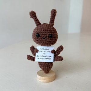 Könnte beinhalten: Gehäkelte braune Ameisenfigur mit schwarzen Augen und einem lächelnden Mund. Die Ameise hält ein Schild mit der Aufschrift "Positive Ant" und einer motivierenden Botschaft. Die Figur steht auf einem kleinen Holzsockel.