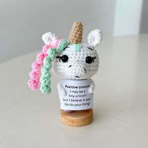 Puede incluir: Figura de unicornio de ganchillo con cuerpo blanco, cuerno marrón y melena rosa y verde. El unicornio sostiene un cartel que dice "Positive Unicorn. I may be a tiny unicorn but I believe in you. Go do your thing!"