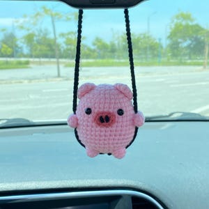 May include: A pink crocheted pig car charm with black eyes, a dark pink snout, and small arms and legs. The pig is suspended by a black cord, hanging from a car's rearview mirror. The charm is a cute, handmade accessory.
