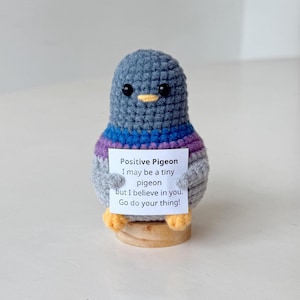 May include: A crocheted pigeon figurine in shades of gray, purple, and blue, holding a small sign. The sign reads "Positive Pigeon" and includes an inspirational message. The pigeon has a yellow beak and small wooden feet.