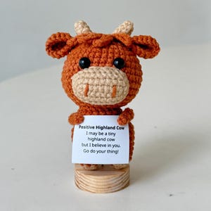Könnte beinhalten: Gehäkelte Highland-Kuh-Figur in Braun- und Beigetönen, die ein Schild hält. Auf dem Schild steht "Positive Highland Cow" mit einer inspirierenden Botschaft. Die Kuh ist auf einem kleinen Holzsockel befestigt.