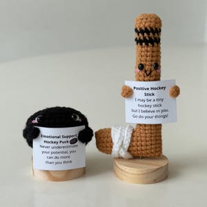 May include: Two crocheted stuffed toys, a black hockey puck and a brown hockey stick, each holding a sign with encouraging messages. The hockey puck says "Emotional Support Hockey Puck Never underestimate your potential, you can do more than you think." The hockey stick says "Positive Hockey Stick I may be a tiny hockey stick but I believe in you. Go do your things!"