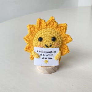 May include: A crocheted yellow sun with a smiling face holds a sign that reads "A little sunshine to brighten your day." The sun is mounted on a small wooden base. The sign also features a small sun illustration.