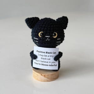 Könnte beinhalten: Gehäkelte schwarze Katzenfigur mit gelben Augen und weißen Schnurrhaaren. Die Katze hält ein Schild mit dem Text "Positive Black Cat, I may be a tiny black cat but I believe in you. You're Meow-nderful."