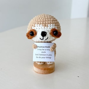 May include: A crocheted sloth figurine with a light brown head, white face, and dark brown eyes. The sloth holds a sign that reads "Positive Sloth, I may be a tiny sloth but I believe in you. Go do your thing!" The figurine sits on a wooden base.