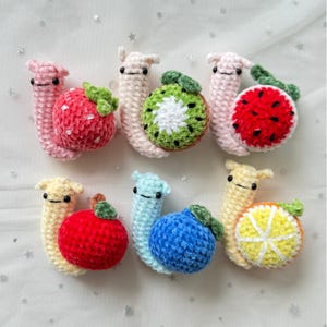 Puede incluir: Seis caracoles de crochet con diferentes conchas de frutas y verduras. Los caracoles son rosas, amarillos, azul claro, verdes y blancos. Las conchas son fresa, manzana, arándano, kiwi, sandía y naranja.