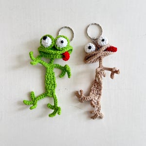 Könnte beinhalten: Zwei Frosch-Schlüsselanhänger aus Häkelarbeit. Einer ist grün mit weißen und schwarzen Augen und einer roten Zunge. Der andere ist hellbraun mit weißen und schwarzen Augen und einer roten Zunge. Beide haben silberne Schlüsselringe.