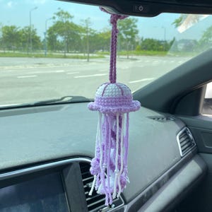 May include: A handmade crochet jellyfish car charm in shades of purple and white. The jellyfish has a round, domed head with a frilly edge and long, dangling tentacles. It is suspended from a purple cord, designed to hang from a car's rearview mirror.