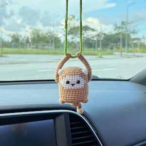 May include: A handmade crochet monkey car charm in shades of brown and white, hanging from a green string. The monkey has a smiling face and is suspended inside a car.