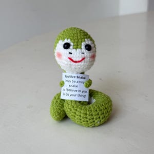May include: A small, green, crocheted snake toy with a white face, black bead eyes, and pink cheeks. The snake holds a white sign that reads: Positive Snake, I may be a tiny snake but I believe in you. Go do your thing! The body is coiled.