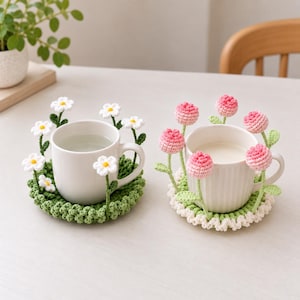 Puede incluir: Dos tazas de cerámica blanca sobre posavasos redondos de ganchillo. El posavasos izquierdo es verde con margaritas blancas en tallos. El derecho es blanco con capullos de flores rosas en tallos verdes. Cada posavasos tiene detalles florales.