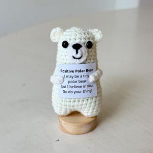 May include: A small white crocheted polar bear figurine with black bead eyes and a stitched nose. The bear holds a white sign that reads: Positive Polar Bear, I may be a tiny polar bear but I believe in you. Go do your thing! It stands on a base.