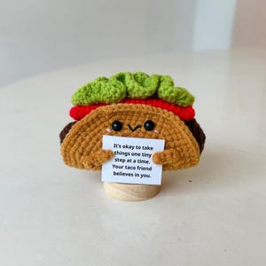 May include: A crocheted taco with a smiling face, green lettuce, and red tomato. The taco holds a small sign that reads, "It's okay to take things one tiny step at a time. Your taco friend believes in you."