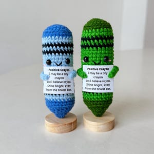Pode incluir: Dois lápis de crochê feitos à mão, um azul e outro verde, cada um com um rosto sorridente e segurando uma pequena placa. As placas dizem "Lápis Positivo" e uma mensagem motivacional. Eles são exibidos em pequenas bases de madeira.