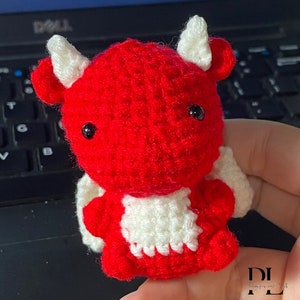 Puede incluir: Un drag&oacute;n rojo de crochet con alas blancas y ojos negros. El drag&oacute;n est&aacute; sentado en una mano.