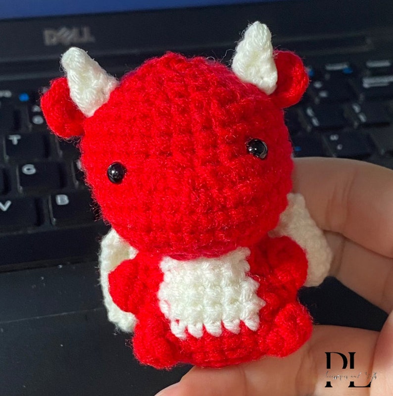 Puede incluir: Un drag&oacute;n rojo de crochet con alas blancas y ojos negros. El drag&oacute;n est&aacute; sentado en una mano.