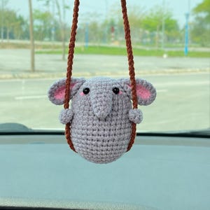 May include: A crocheted elephant car charm in gray with pink inner ears and brown hanging cords. The elephant has small black eyes and a trunk. The charm is hanging from a car's rearview mirror.