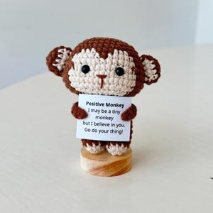 Puede incluir: Figura de mono de ganchillo en marrón y crema, sosteniendo un letrero que dice "Positive Monkey I may be a tiny monkey but I believe in you. Go do your thing!". El mono está sobre una base de madera.