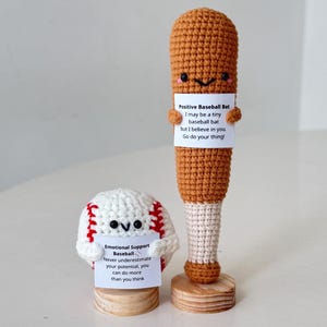 Puede incluir: Dos artículos de béisbol de ganchillo: una pelota y un bate. La pelota es blanca con costuras rojas y el texto "Emotional Support Baseball". El bate es marrón con el texto "Positive Baseball Bat".