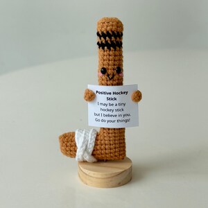 May include: A crocheted hockey stick with a smiling face holding a sign that says "Positive Hockey Stick I may be a tiny hockey stick but I believe in you. Go do your things!"