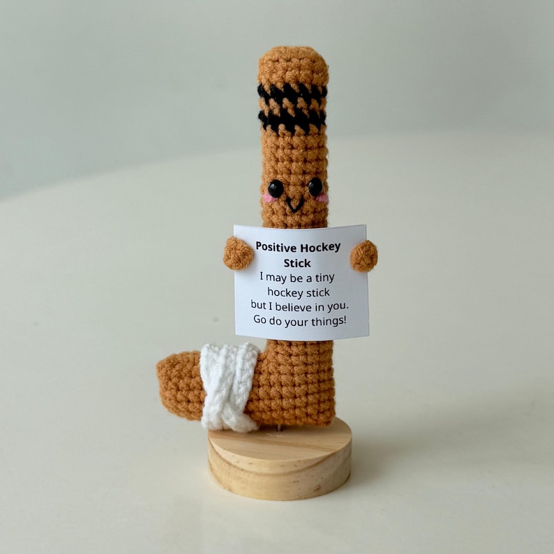 May include: A crocheted hockey stick with a smiling face holding a sign that says "Positive Hockey Stick I may be a tiny hockey stick but I believe in you. Go do your things!"