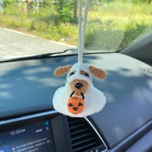 May include: A white and brown crocheted dog car charm with a small orange pumpkin bucket. The dog has brown ears and a black nose. The charm hangs from a white string, perfect for a rearview mirror.