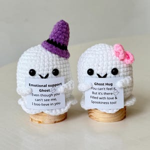 May include: Two handmade crochet ghost figures. One ghost wears a purple witch hat, the other a pink bow. Each holds a sign with a message. The ghosts are white with black eyes and are mounted on wooden bases.