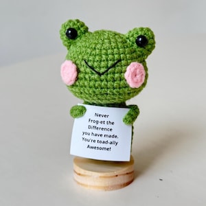 May include: A handmade green crochet frog figurine with pink cheeks and black eyes. The frog holds a white sign with the text "Never Frog-et the Difference you have made. You're toad-ally Awesome!" The frog is mounted on a small wooden base.
