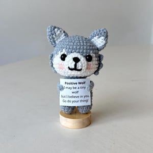 Könnte beinhalten: Eine kleine, grau-weiße gehäkelte Wolfsfigur mit schwarzen Augen und rosa Wangen, die auf einem runden Holzsockel steht. Der Wolf hält ein weißes Schild mit der Aufschrift: Positive Wolf, I may be a tiny wolf but I believe in you.