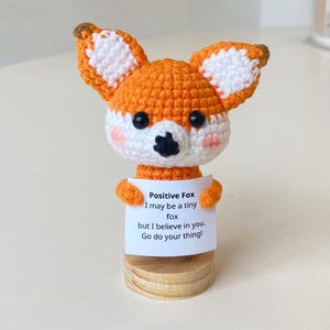 Puede incluir: Un pequeño zorro naranja de crochet sosteniendo un cartel que dice "Positive Fox I may be a tiny fox but I believe in you. Go do your thing!"