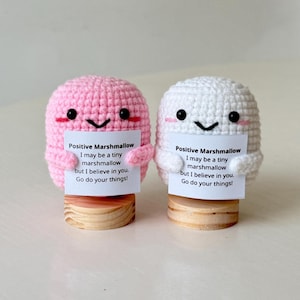 Op de afbeelding: Twee gehaakte marshmallow-figuren, één roze en één wit, die elk een bord vasthouden. Op de borden staat "Positive Marshmallow" en een motiverende boodschap. De figuren staan op houten voetstukken.