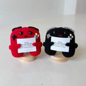 Puede incluir: Dos mandos de juego de ganchillo, uno rojo y otro negro, cada uno sosteniendo una tarjeta blanca con texto. El mando rojo tiene una cruz negra y botones coloridos. El mando negro tiene detalles grises y botones coloridos. El texto dice: "YOU'RE NEXT LEVEL AWESOME."