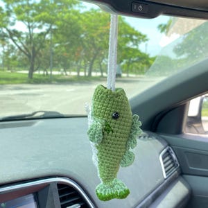 Op de afbeelding: Een gehaakte vis auto-hanger in groen en wit, hangend aan een wit touwtje. De handgemaakte vis heeft zwarte ogen en gedetailleerde vinnen. De hanger hangt in een auto, met een vage buitenomgeving op de achtergrond.