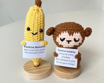Peut inclure: Deux peluches en crochet, une banane et un singe, tenant des pancartes avec des messages positifs. La banane dit "Positive Banana When you focus good The good gets better". Le singe dit "Positive monkey I may be a tiny monkey but I believe in you. Go do your things!"