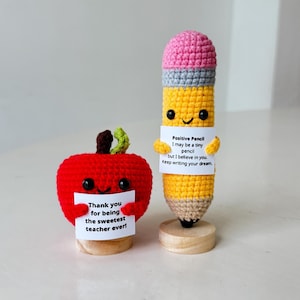 Puede incluir: Figuras de ganchillo de manzana y lápiz. La manzana roja sostiene un letrero que dice: "¡Gracias por ser el profesor más dulce!" El lápiz amarillo sostiene un letrero que dice: "Lápiz positivo Puede que sea un lápiz pequeño, pero creo en ti. Sigue escribiendo tu sueño."
