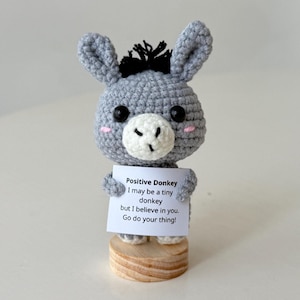 May include: A gray crocheted donkey figurine with black eyes, a white muzzle, and pink cheeks. The donkey holds a sign that reads "Positive Donkey I may be a tiny donkey but I believe in you. Go do your thing!" The figurine sits on a wooden base.