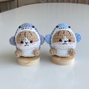 May include: Two crocheted plush toys shaped like a shark wearing a white hooded sweater. The shark toys are standing on wooden bases. One shark is light blue and the other is purple.