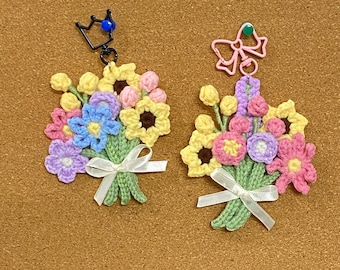 Könnte beinhalten: Zwei Schlüsselanhänger mit gehäkelten Blumensträußen. Einer hat einen schwarzen Kronenanhänger mit einer blauen Perle, der andere einen rosa Schleifenanhänger. Die Sträuße zeigen gelbe, rosa, blaue und lila Blumen, die mit weißen Bändern gebunden sind.