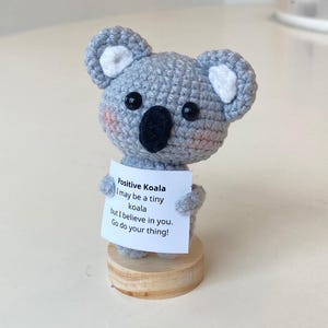 Könnte beinhalten: Ein kleiner grauer Häkel-Koala, der ein Schild mit der Aufschrift "Positiver Koala Ich bin vielleicht ein kleiner Koala, aber ich glaube an dich. Mach dein Ding!" hält.