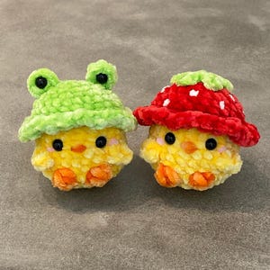 Può includere: Due pulcini gialli all'uncinetto con cappelli diversi. Un pulcino indossa un cappello di rana verde e l'altro indossa un cappello di fragola rosso e bianco.