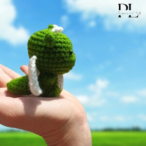 Puede incluir: Un peque&ntilde;o drag&oacute;n verde de crochet con alas blancas, sostenido en una mano contra un cielo azul con nubes blancas.