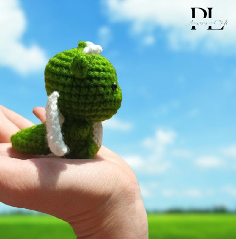 Puede incluir: Un peque&ntilde;o drag&oacute;n verde de crochet con alas blancas, sostenido en una mano contra un cielo azul con nubes blancas.