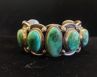 May include: A silver cuff bracelet with five oval turquoise stones set in a silver bezel. The stones are a mix of green and blue hues.