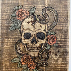 May include: A wood burned image of a skull with a snake wrapped around it. The skull is surrounded by roses and leaves. The image is on a wooden background.