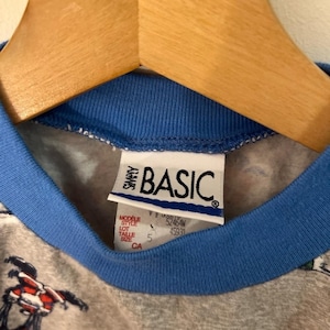 May include: Close-up of a gray t-shirt with a blue collar and the tag "SIMPLY BASIC." The tag also includes the size "5" and the letters "CA." The shirt is hanging on a wooden hanger.