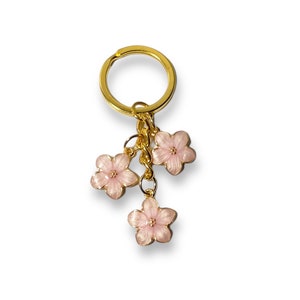 May include: A gold key chain with three pink flower charms. The flowers have a gold outline and a light pink center.
