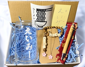 May include: A brown cardboard box filled with blue and white tissue paper. The box contains a clear glass with Japanese characters, a wooden bracelet with Japanese characters, a pair of pink flower earrings, a wooden pen with Japanese characters, and a note with Japanese characters.