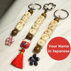 May include: Three wooden keychains with Japanese characters, each with a different charm: a pink flower, a red and white daruma doll, and a blue flower.