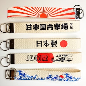 May include: Five different keychains with Japanese designs. The keychains are made of beige fabric with black and red printed designs. The designs include a rising sun, Japanese text, a red circle, a car, and a wave pattern.